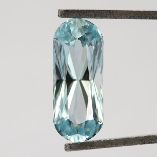 Light blue long-oval sapphire held crown-side toward the camera in metal tweezers, with an elongated table and rounded ends.