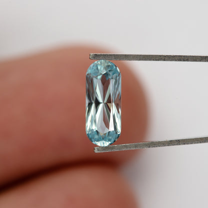 Light blue sapphire in tweezers against a fingertip, showing its small scale.