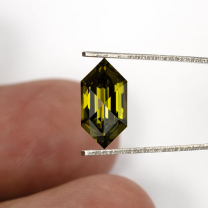 Yellow-green lab-grown sapphire held in tweezers in front of fingertips to show its size.