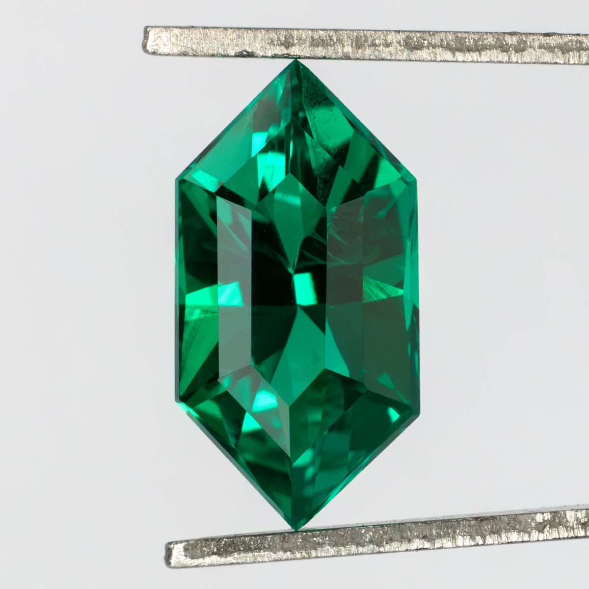 Face-up green lab-grown emerald held vertically in tweezers, showing its crown facets and fine visible internal features.