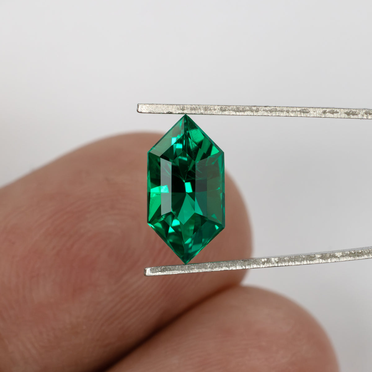 Green lab-grown emerald held in tweezers in front of fingertips to show its size and outline.