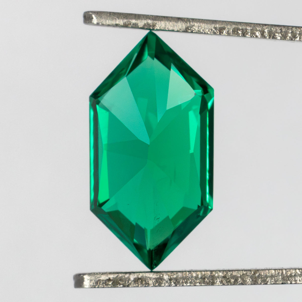 Back of the green lab-grown emerald held vertically in tweezers, showing broad pavilion facets.