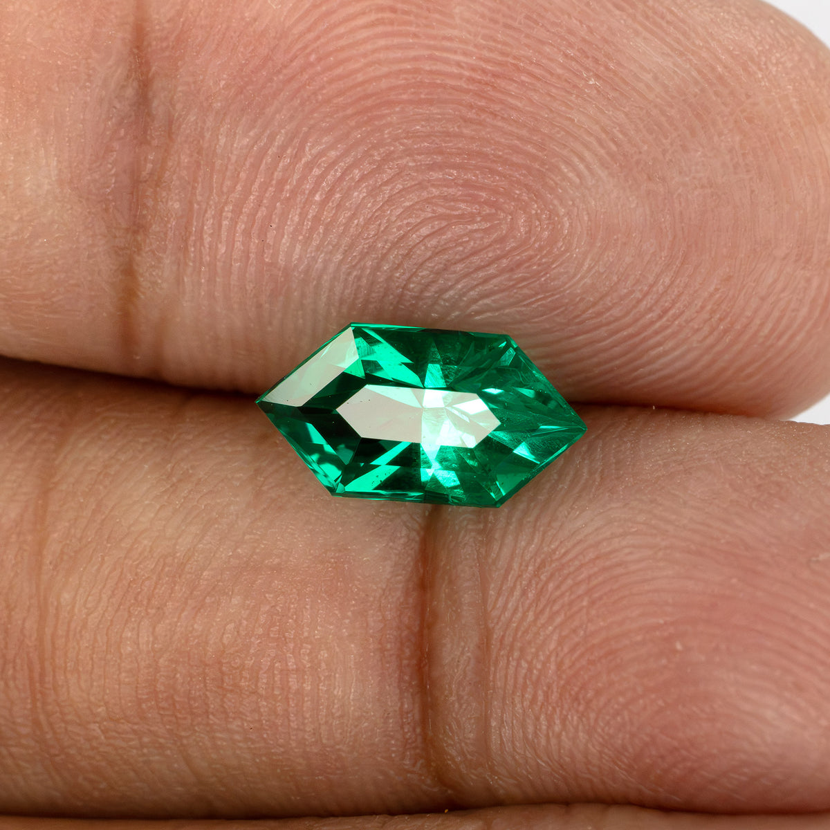 The lab-grown emerald on fingertips at a tilted crown angle, with a broad pale reflection across its center.