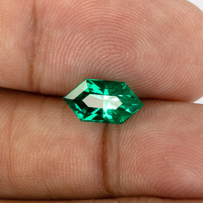 The lab-grown emerald on fingertips at a tilted crown angle, with a broad pale reflection across its center.