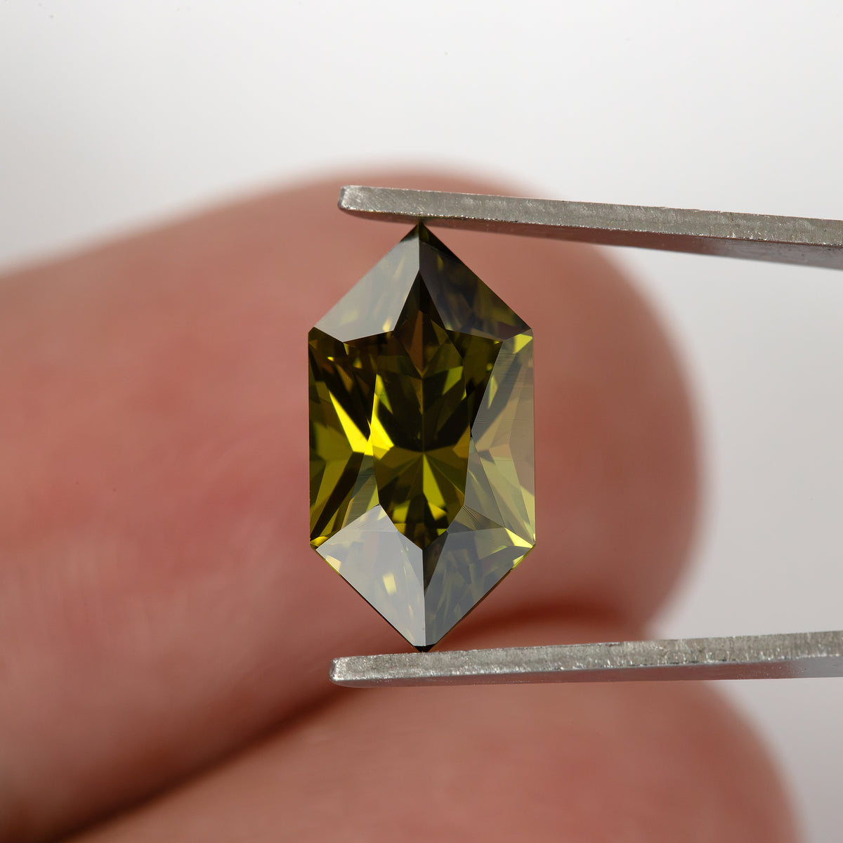 Face-up yellow-green sapphire in tweezers against a fingertip for scale.