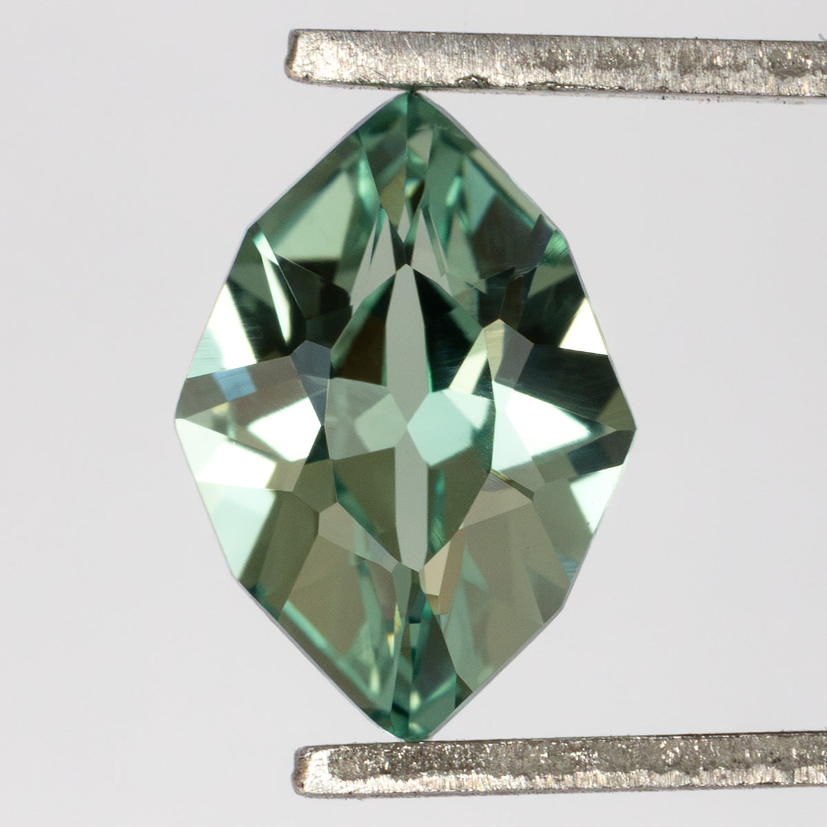 Face-up green lab-grown sapphire held vertically in tweezers, showing its pointed outline and crown facets.