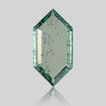 Portrait cut elongated hexagonal green gemstone showing internal linear inclusions and reflection on grey background.