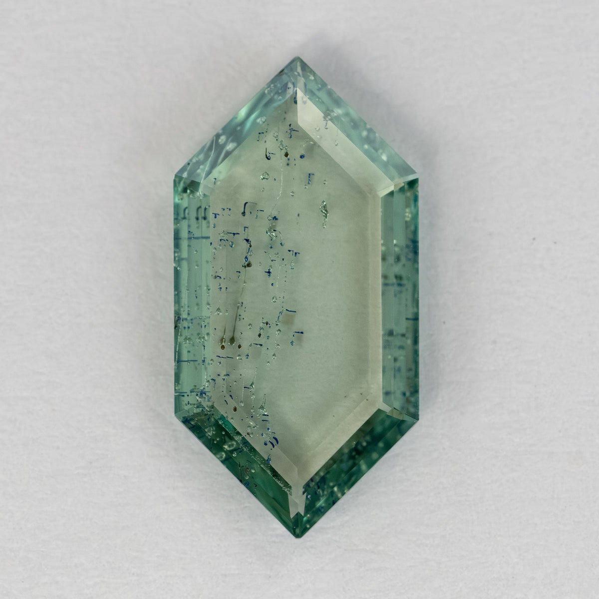 Elongated hexagonal green gemstone lying flat on white textured surface showing transparent body and inclusions.