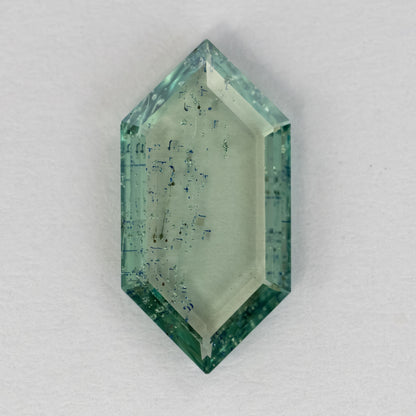 Elongated hexagonal green gemstone lying flat on white textured surface showing transparent body and inclusions.
