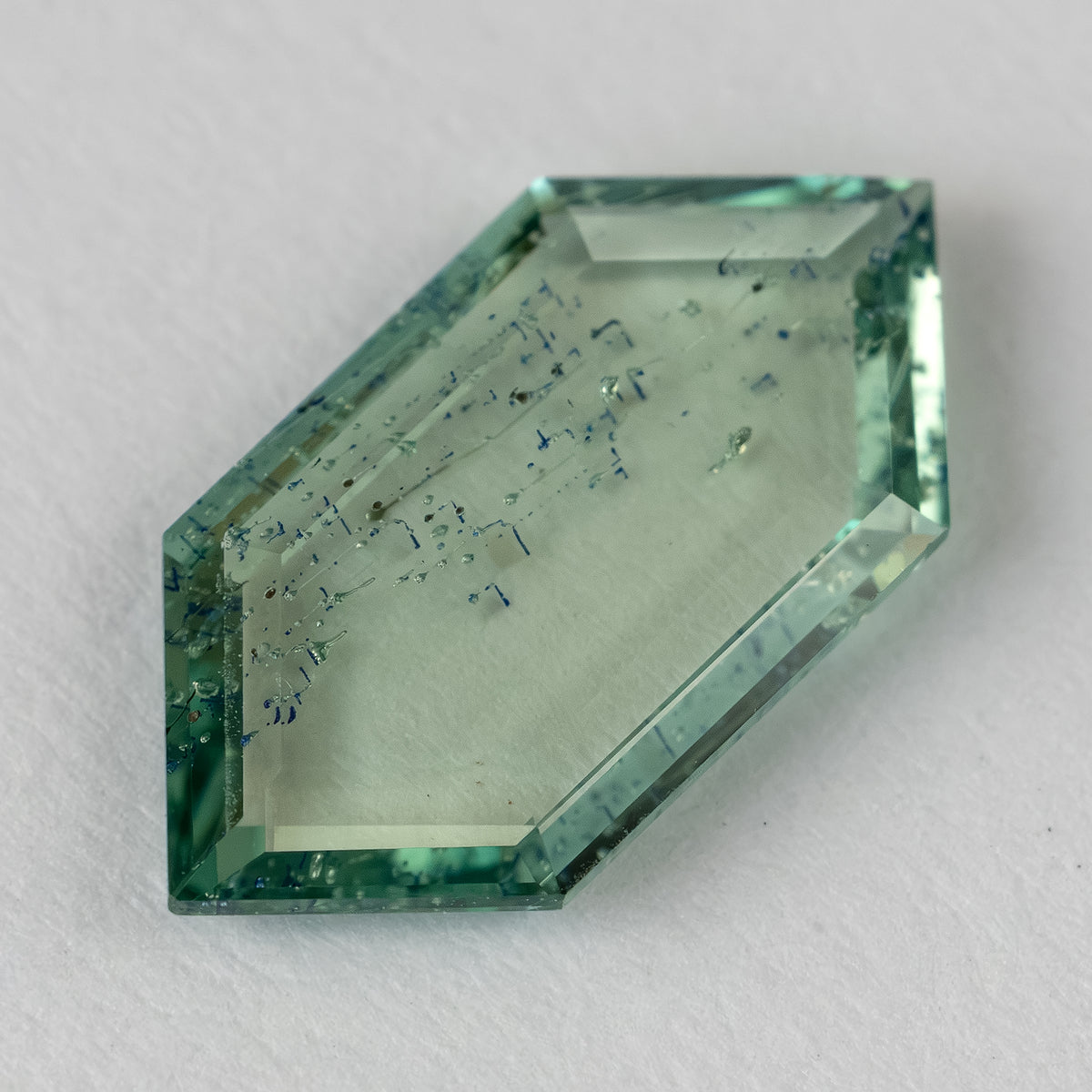 Angled perspective of thin hexagonal green gemstone showing faceted bevel edges and internal patterns on white paper.