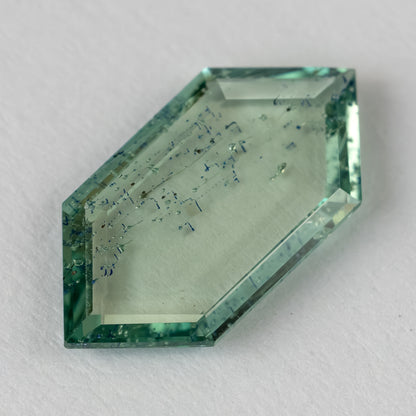 Angled perspective of thin hexagonal green gemstone showing faceted bevel edges and internal patterns on white paper.