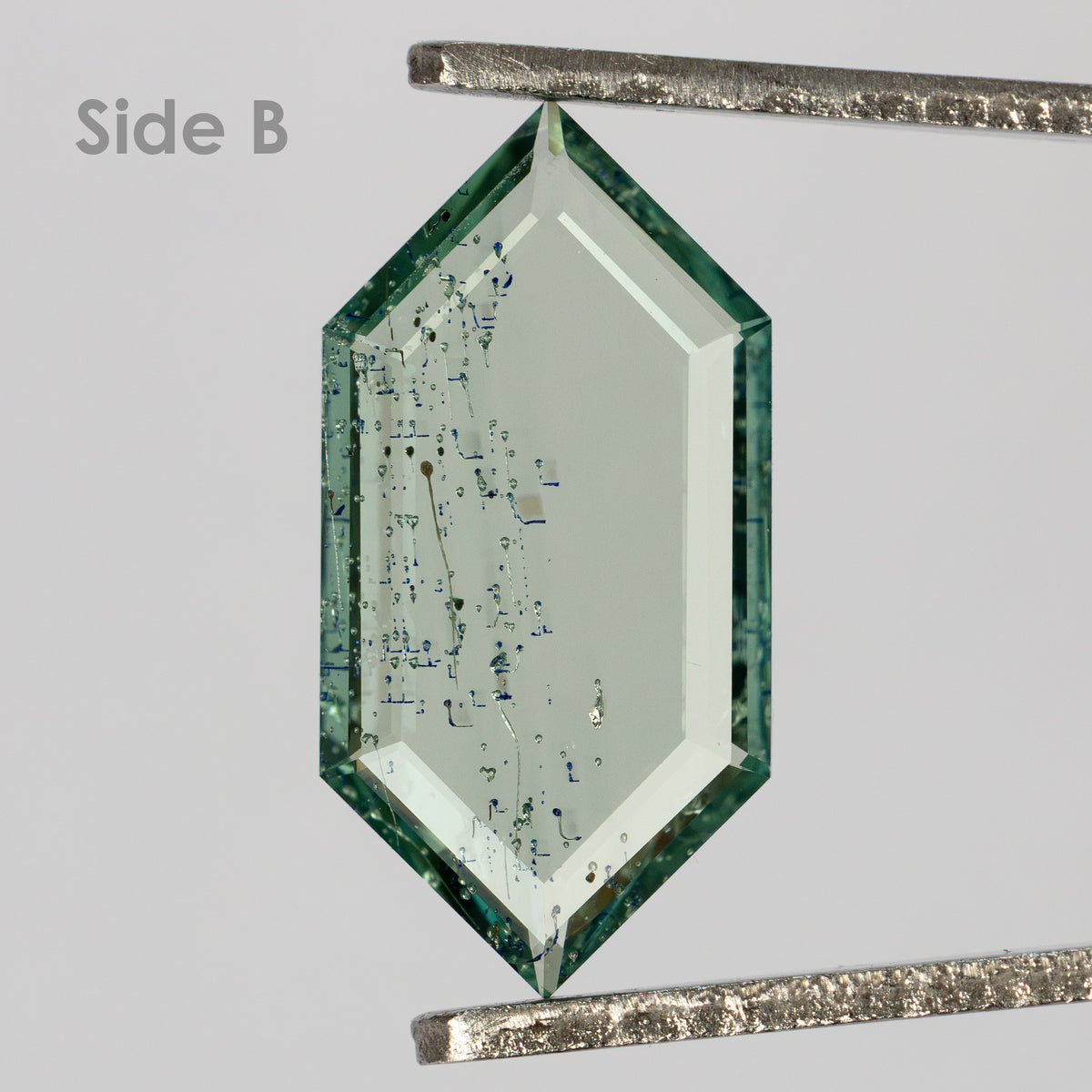 Opposite side of elongated hexagonal green gemstone in tweezers labeled Side B against grey background.