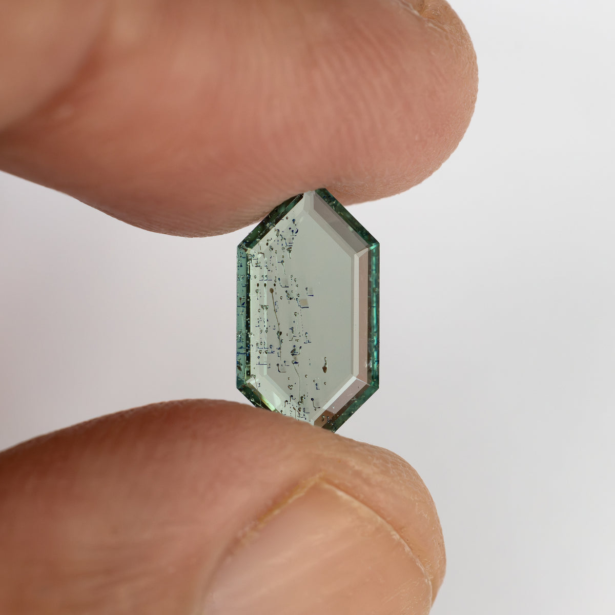 Elongated hexagonal green gemstone held upright between thumb and finger against neutral background.