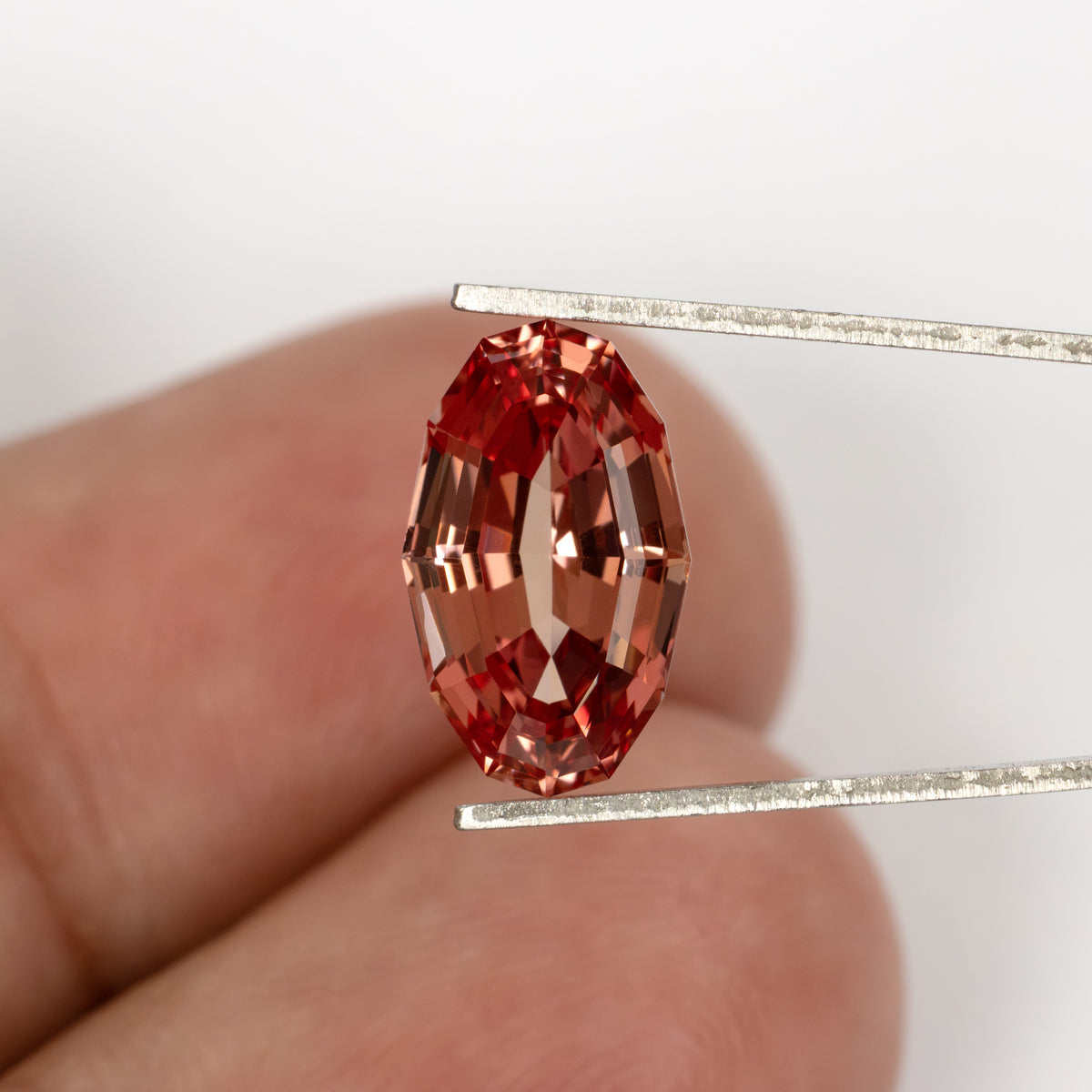 Orange-pink oval lab-grown sapphire held in tweezers in front of fingers for approximate scale.