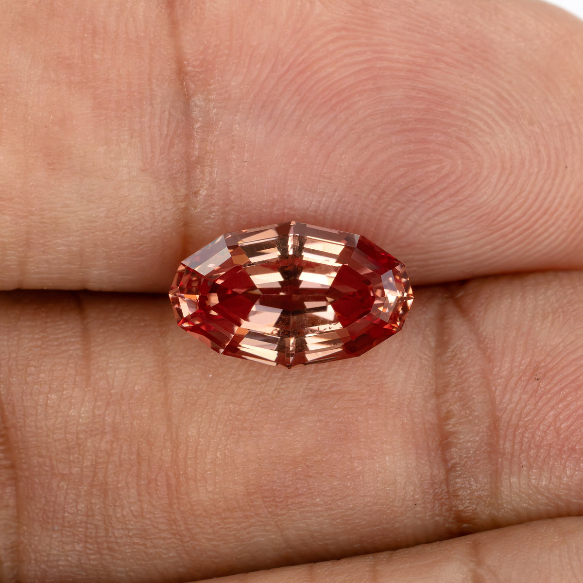 Face-up orange-pink lab-grown sapphire resting on fingers, showing its elongated oval outline and broad step facets.