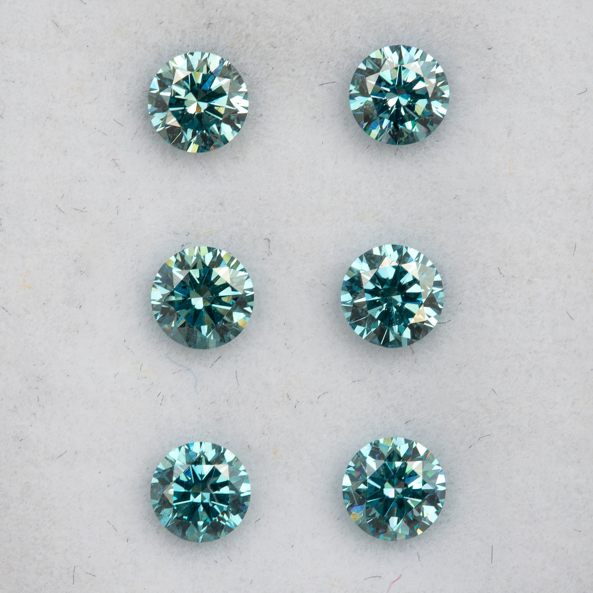 Six round blue diamonds with teal tones and bright facet reflections, arranged face-up in two columns on white padding.