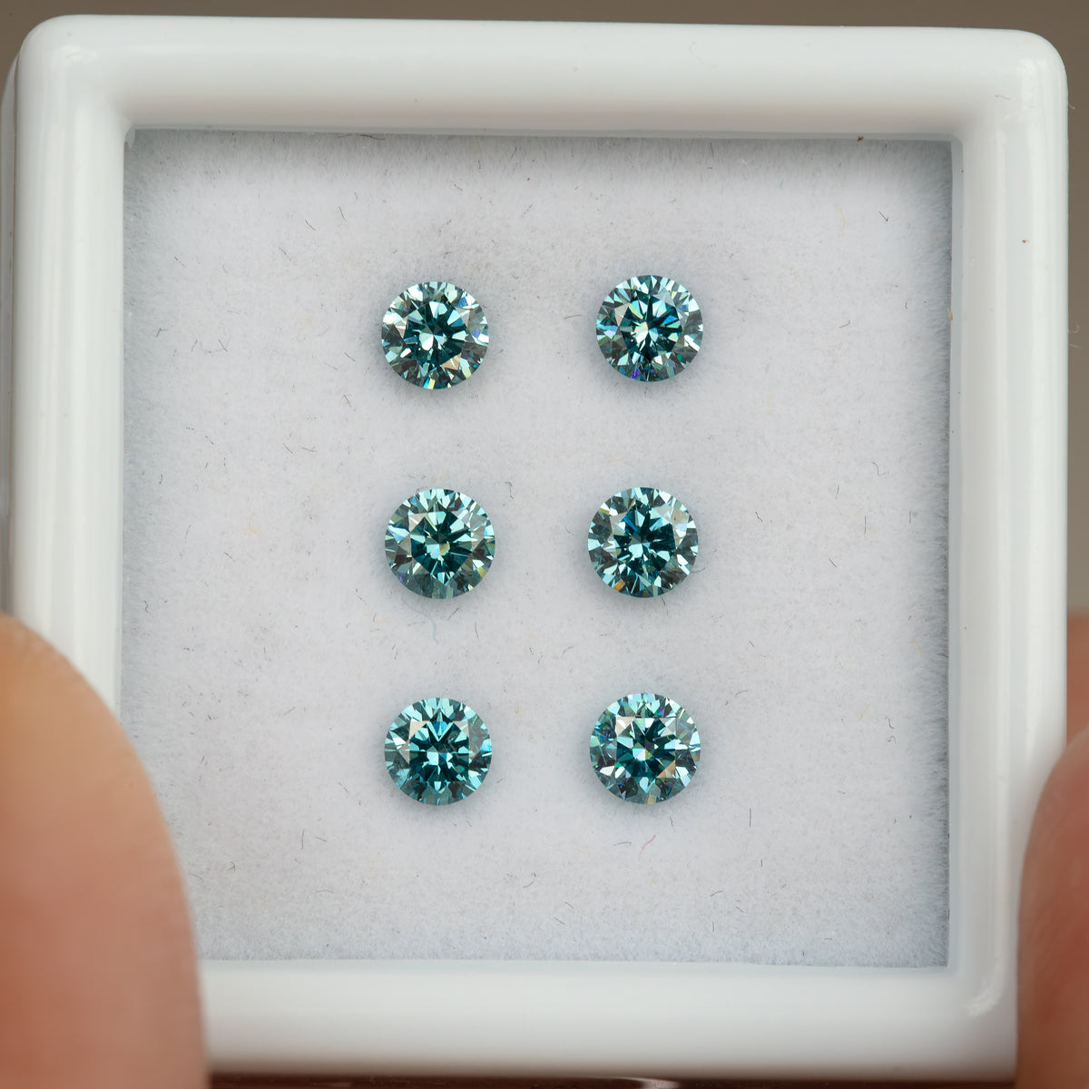 Closer face-up view of six round blue-teal diamonds in white padding, with the display-box rim and fingers visible at the sides.