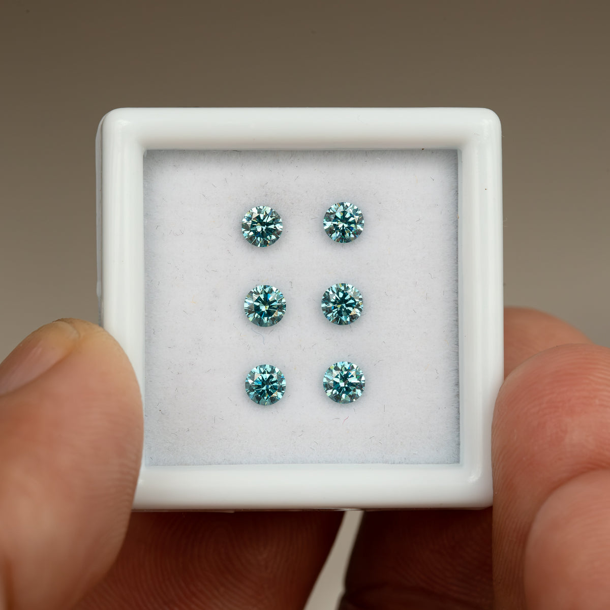 Six round blue-teal diamonds in a square white display box held between fingers, showing the full box for relative scale.