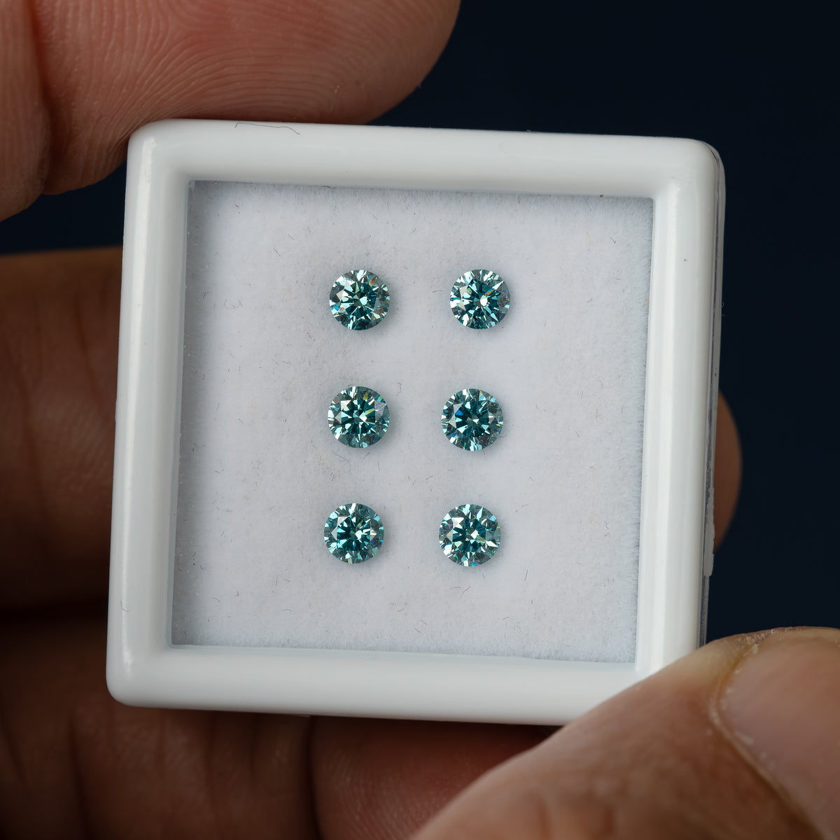 Six round blue-teal diamonds in a slightly tilted white display box held by fingers against a dark background.