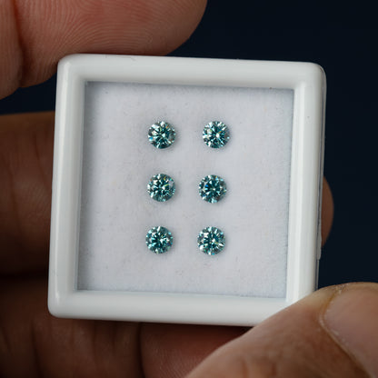 Six round blue-teal diamonds in a slightly tilted white display box held by fingers against a dark background.