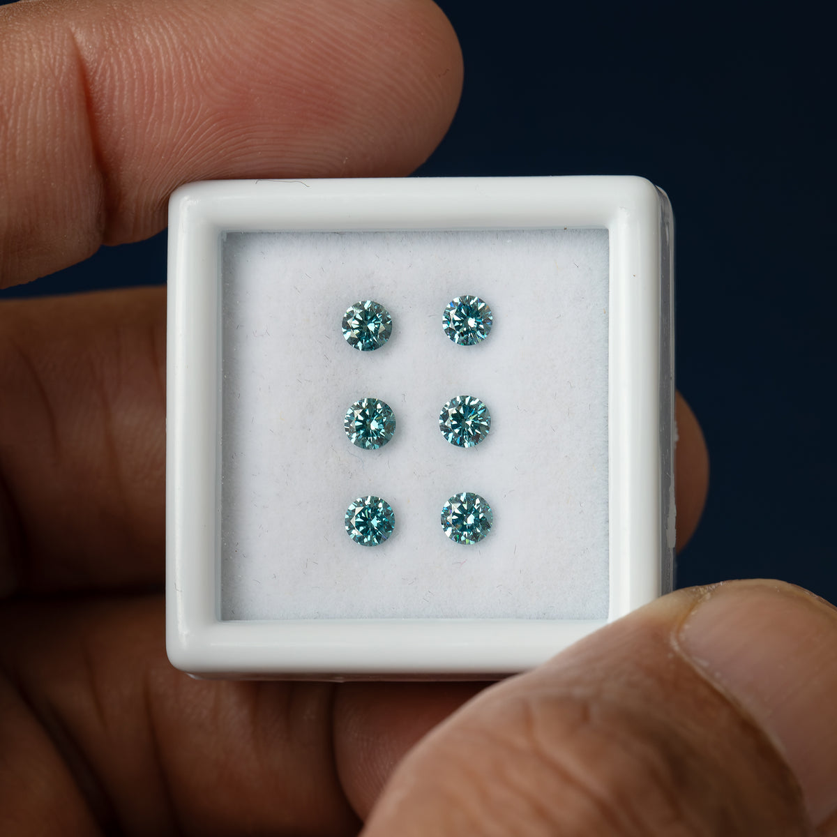 Wider held-box view of six round blue-teal diamonds, with darker centers and bright pale facet reflections against white padding.