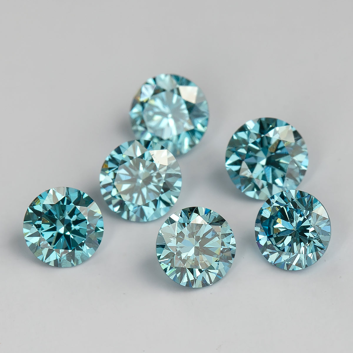 Alternate face-up view of six loose round blue diamonds on a pale surface, showing lighter blue reflections and differences in brightness.