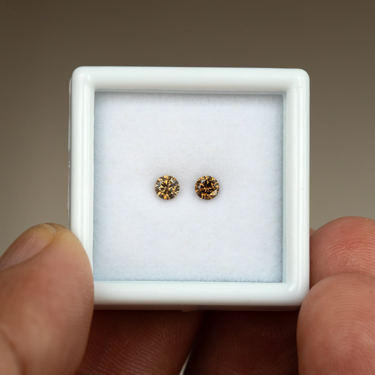 Two round cognac-brown diamonds in a square white display box held between fingers, showing the full box for relative scale.