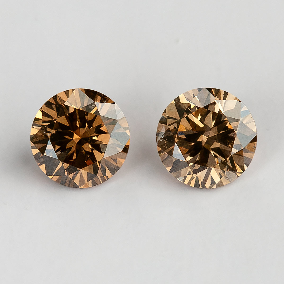 Close face-up view of two round cognac-brown diamonds side by side on a pale surface, showing amber-gold reflections and darker centers.