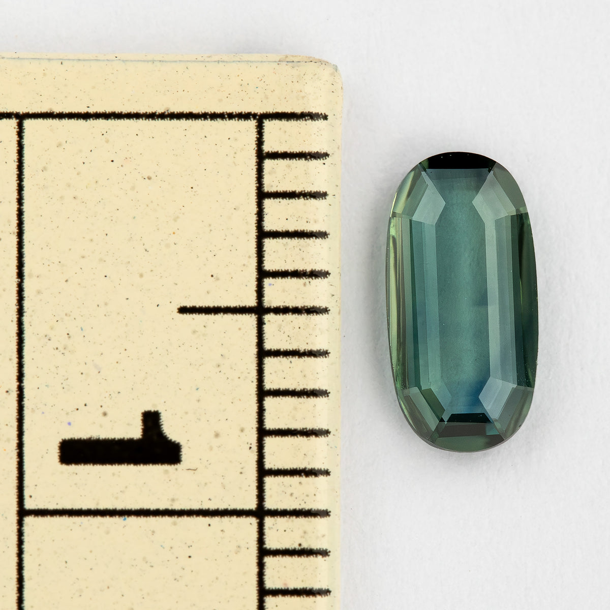 The slim green-blue oval sapphire beside a measurement scale, showing its small physical size.