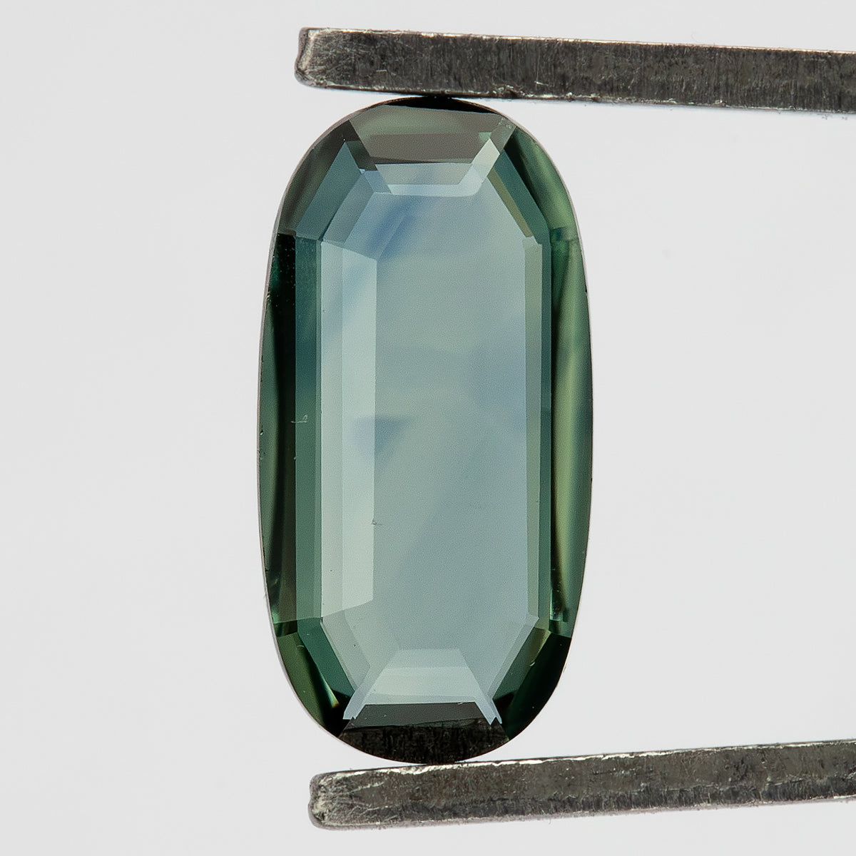 Close-up of the oval sapphire in tweezers, with a transparent center and small features visible near its border.
