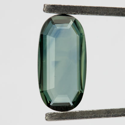 Close-up of the oval sapphire in tweezers, with a transparent center and small features visible near its border.