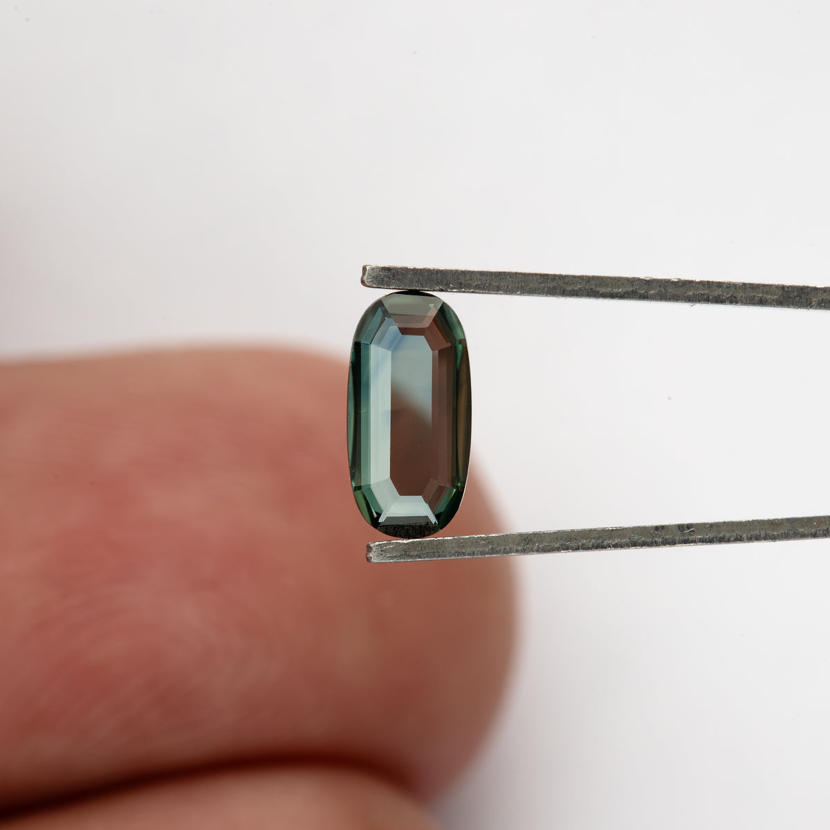Slim oval sapphire held in tweezers in front of a fingertip, showing realistic scale.