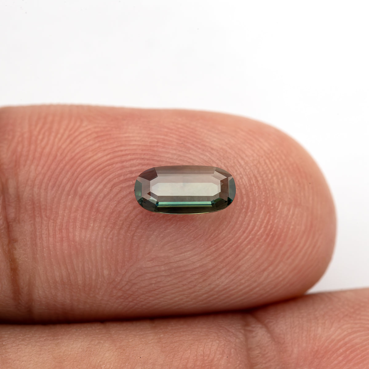Small oval portrait-cut sapphire resting on a fingertip, with the skin visible through the center.