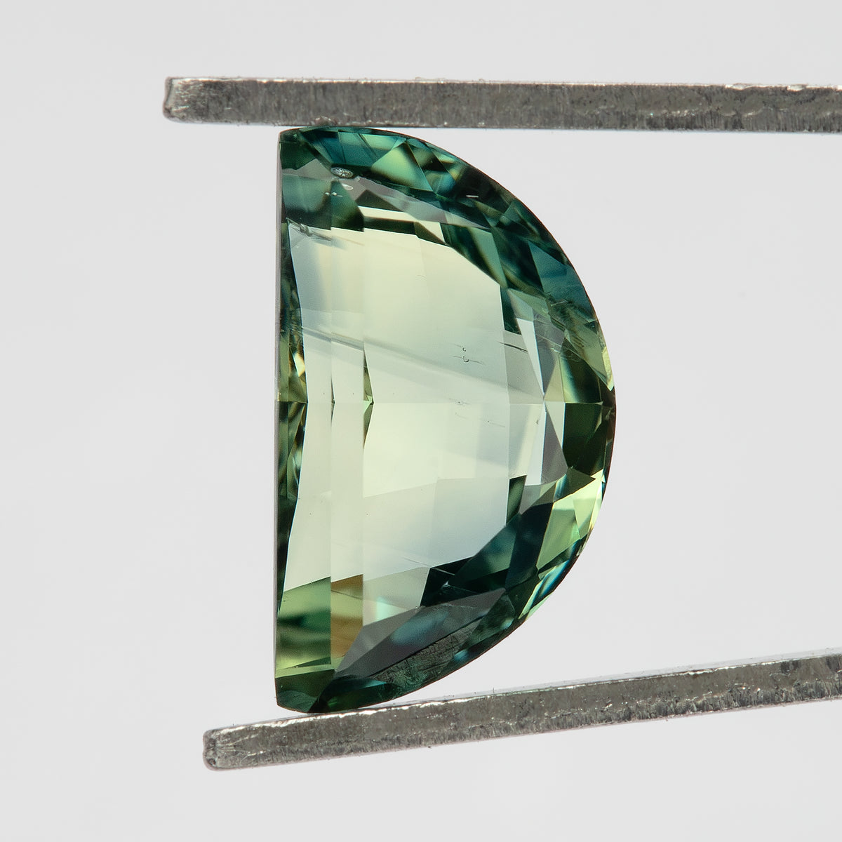 Close face-up view of a half-moon sapphire held vertically in tweezers, showing dark teal-green rim reflections and a pale yellow-green central table.