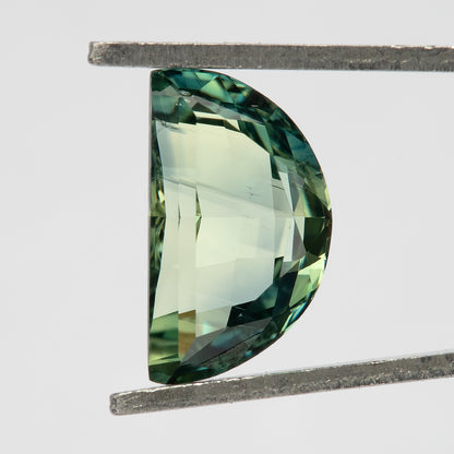 Close face-up view of a half-moon sapphire held vertically in tweezers, showing dark teal-green rim reflections and a pale yellow-green central table.