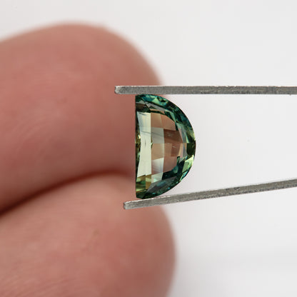 Half-moon sapphire held vertically in tweezers in front of fingers for scale; its curved edge shows teal-green reflections and the finger background is visible through the centre.
