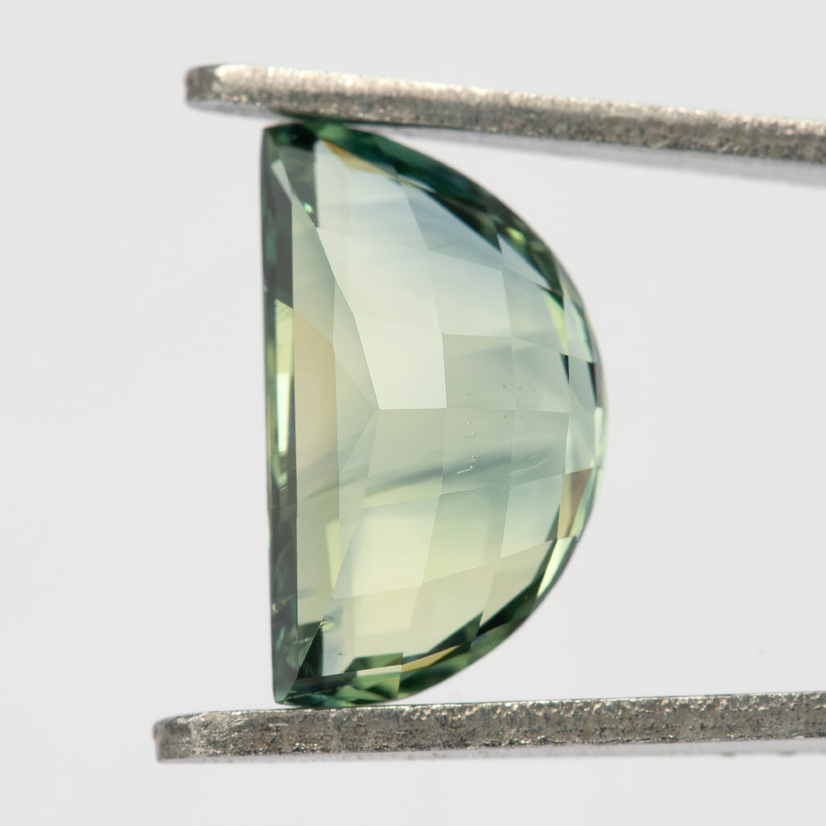 Pavilion-side view of the half-moon sapphire held vertically in tweezers, showing pale yellow-green checkerboard facets and a darker green curved edge.