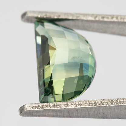 Close tilted pavilion view of the half-moon sapphire in tweezers, with enlarged checkerboard facets reflecting pale green, yellow-green and blue-green tones.