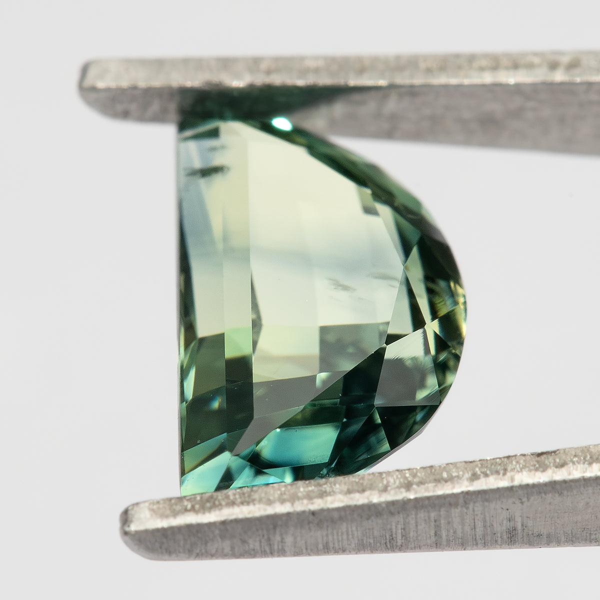 Tilted face-up half-moon sapphire held in tweezers, showing a light yellow-green centre, darker green curved rim and bright blue-green reflections.