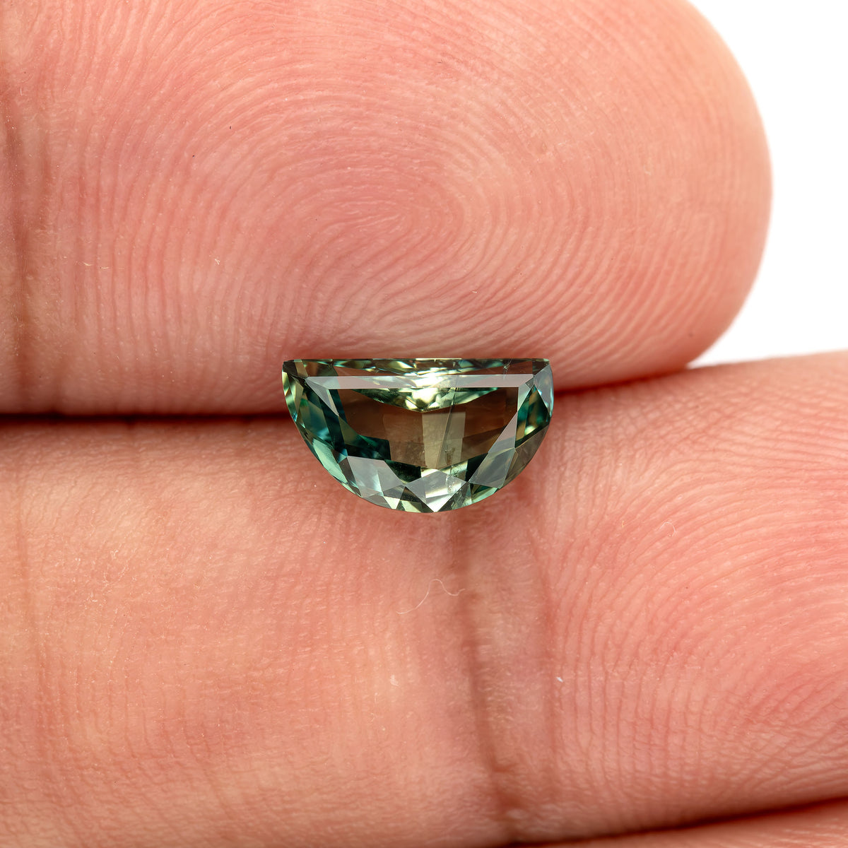 Face-up half-moon sapphire resting on fingers for scale, teal-green rim reflections and the skin background visible through the central table.