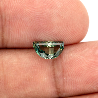 Face-up half-moon sapphire resting on fingers for scale, teal-green rim reflections and the skin background visible through the central table.