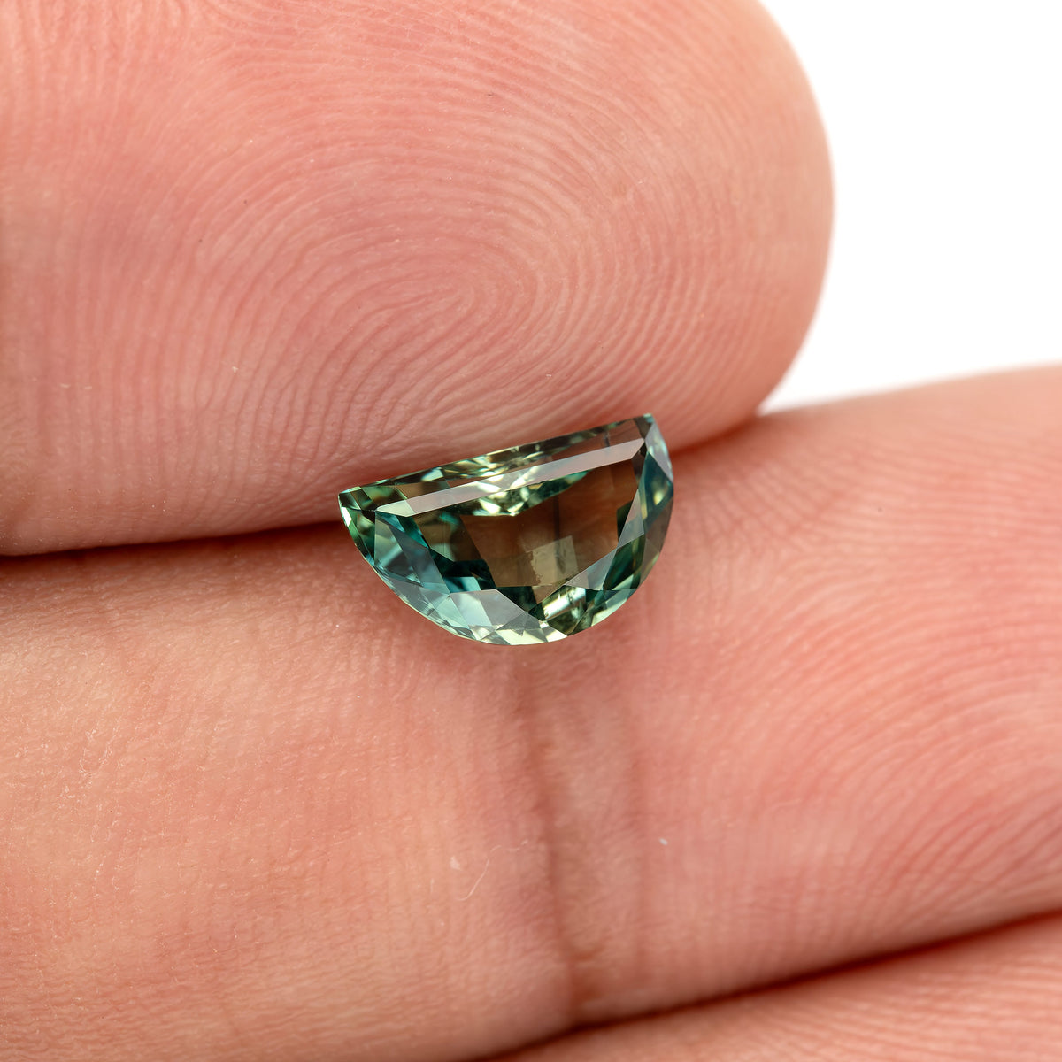 Tilted half-moon sapphire on fingers, showing blue-green edge highlights and the finger background visible through the centre.