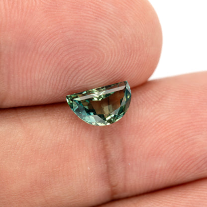 Tilted half-moon sapphire on fingers, showing blue-green edge highlights and the finger background visible through the centre.