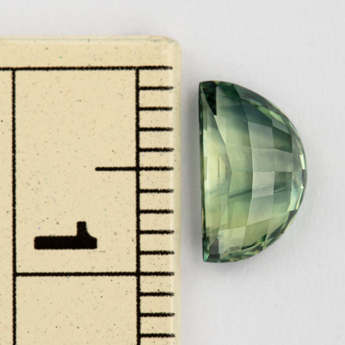 Half-moon sapphire beside a ruler for scale, viewed from the checkerboard pavilion side and pale green and yellow-green facet reflections.