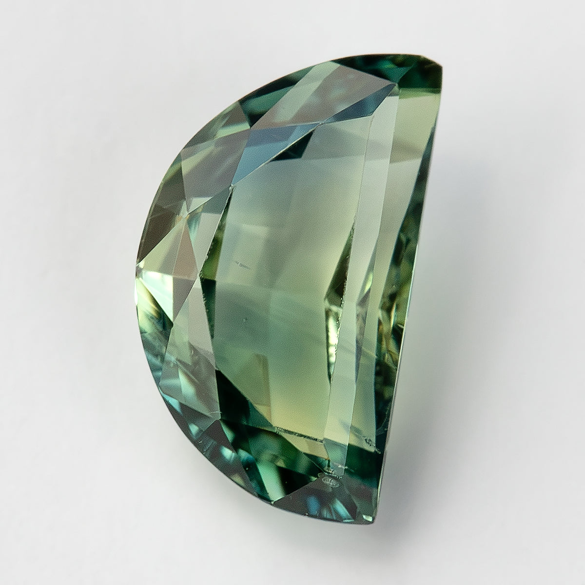Angled face-up view of the loose half-moon sapphire on white, a pale green central table and blue-green highlights around the curved edge.