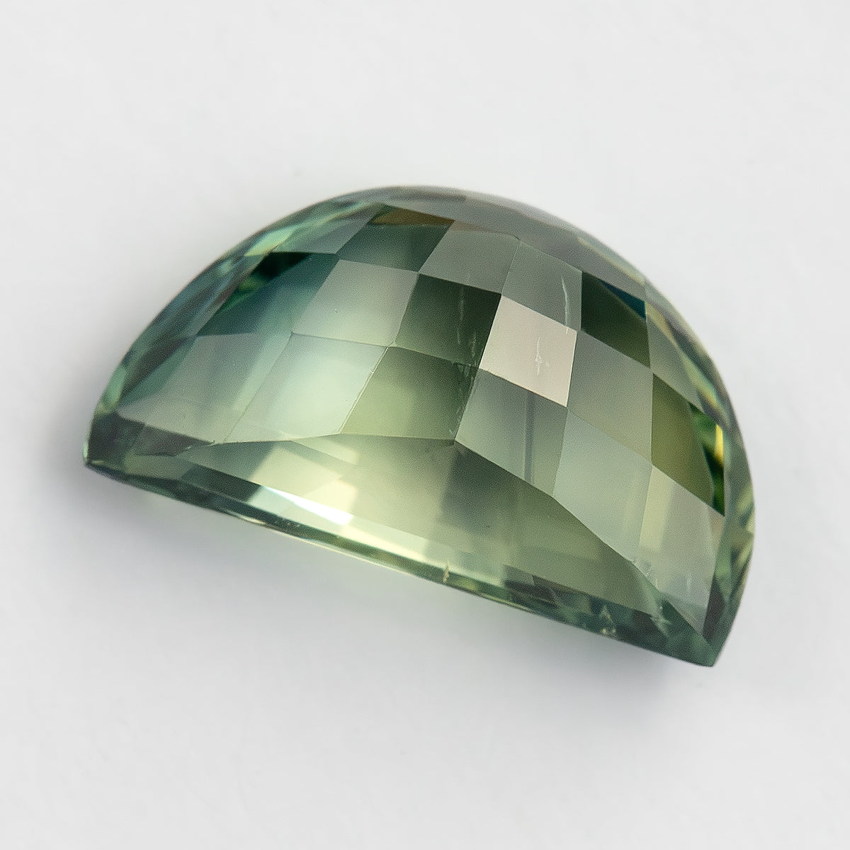 Angled pavilion-side view of the loose half-moon sapphire on white, showing rows of checkerboard facets with soft green, yellow-green and grey reflections.