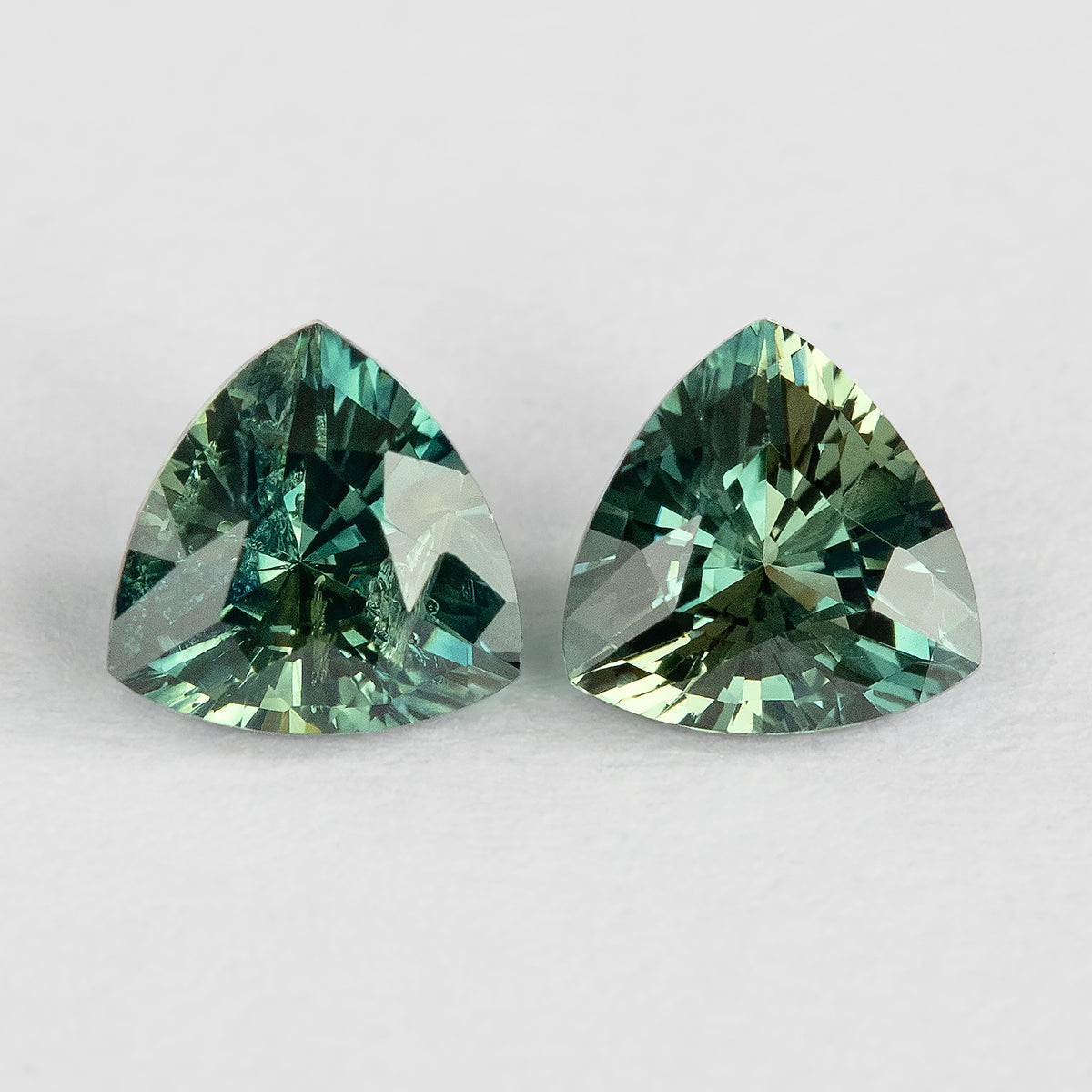 Face-up macro of the two teal-green trillion sapphires on a white surface, showing their outlines, reflections and visible internal characteristics.