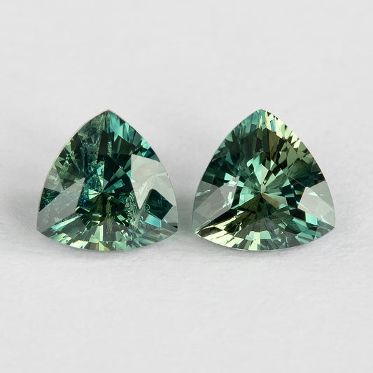 Face-up macro of the two teal-green trillion sapphires on a white surface, showing their outlines, reflections and visible internal characteristics.