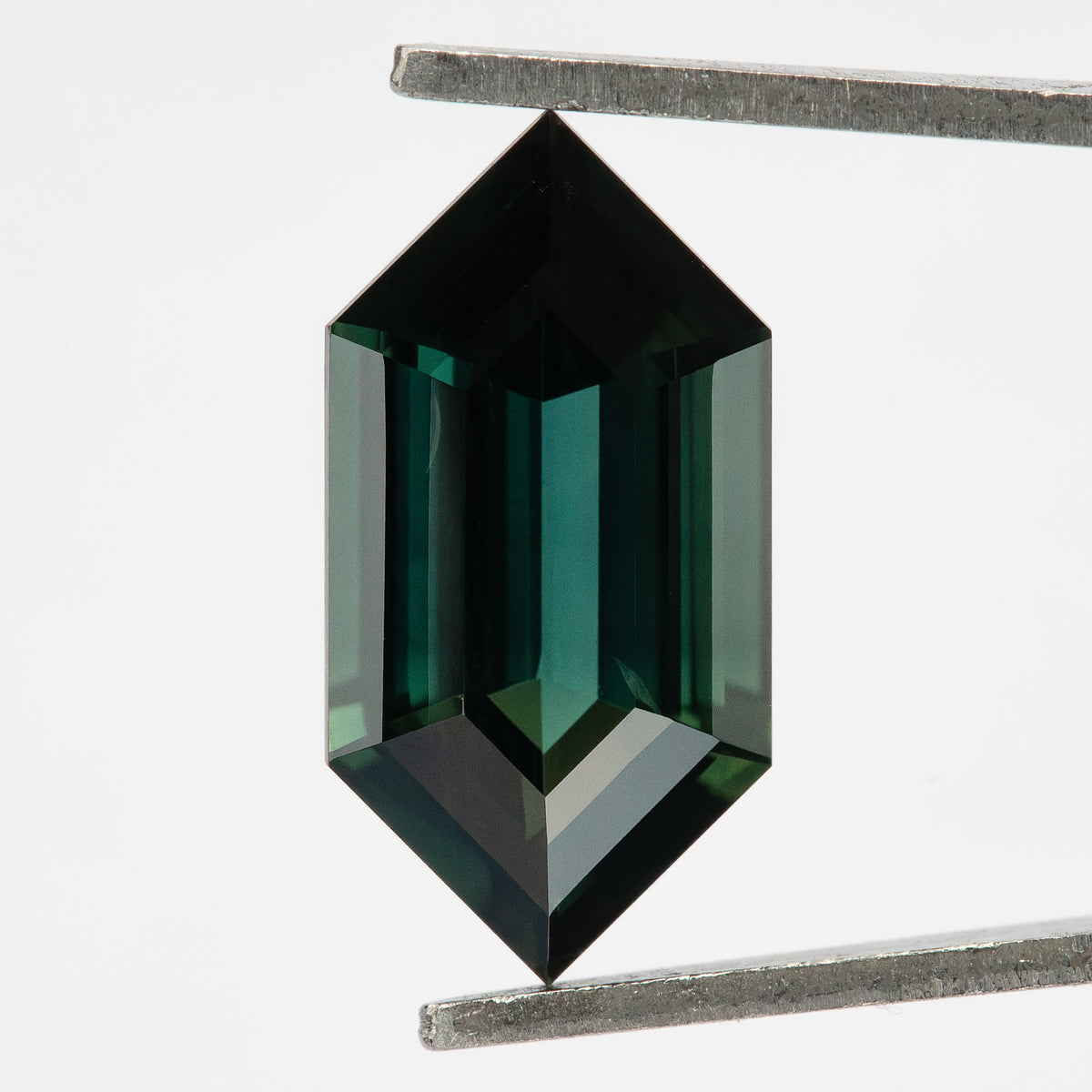 Natural sapphire held vertically in tweezers, showing a darker teal appearance and bright reflections near the lower pointed end.