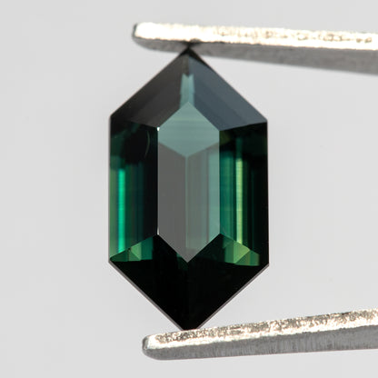 Close face-up view of the sapphire in tweezers, with a pale central facet reflection surrounded by dark teal and bright green bands.
